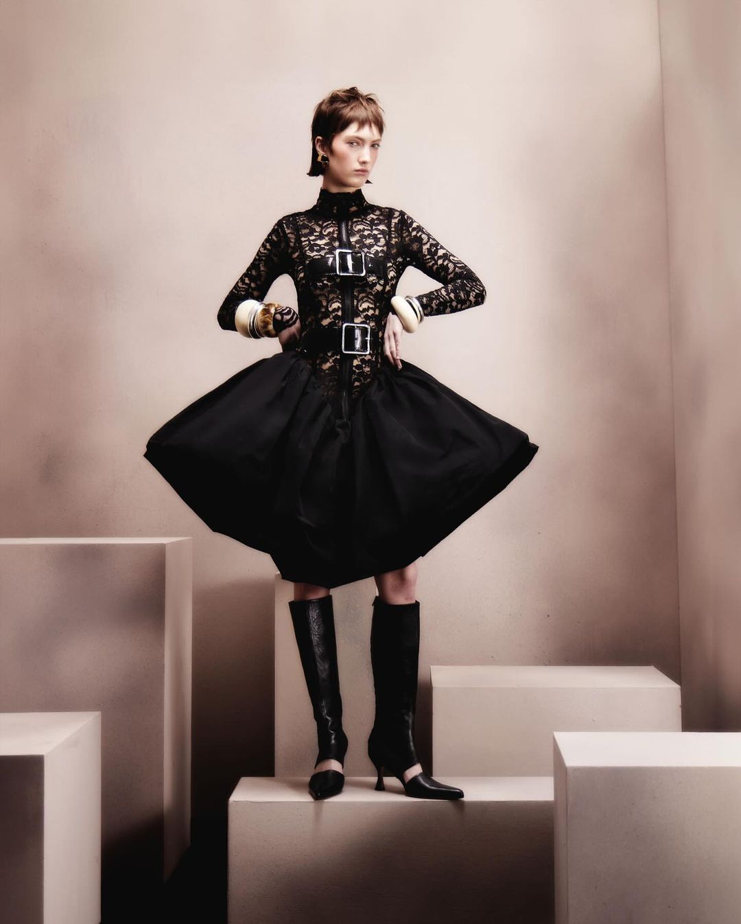 A fashionably dressed adult woman strikes a pose in a black lace-bodice dress, a flared skirt, and knee-high boots, against a neutral backdrop.