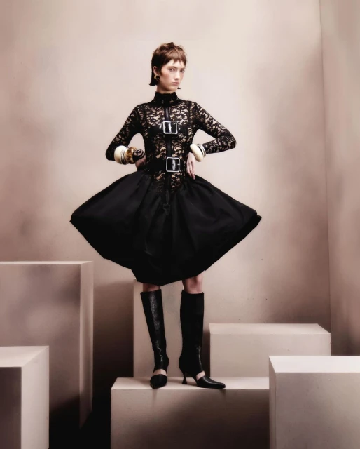 A fashionably dressed adult woman strikes a pose in a black lace-bodice dress, a flared skirt, and knee-high boots, against a neutral backdrop.