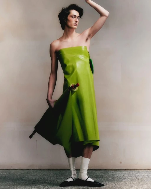 An adult male poses in a green strapless garment, white socks, and black flats in a studio with a neutral-toned wall and concrete floor.