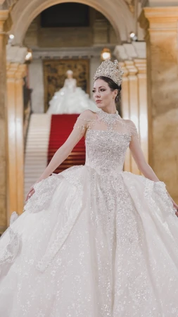 Model wearing an ornate crystal tiara and a voluminous embellished white ballgown on a grand staircase with a red carpet.
