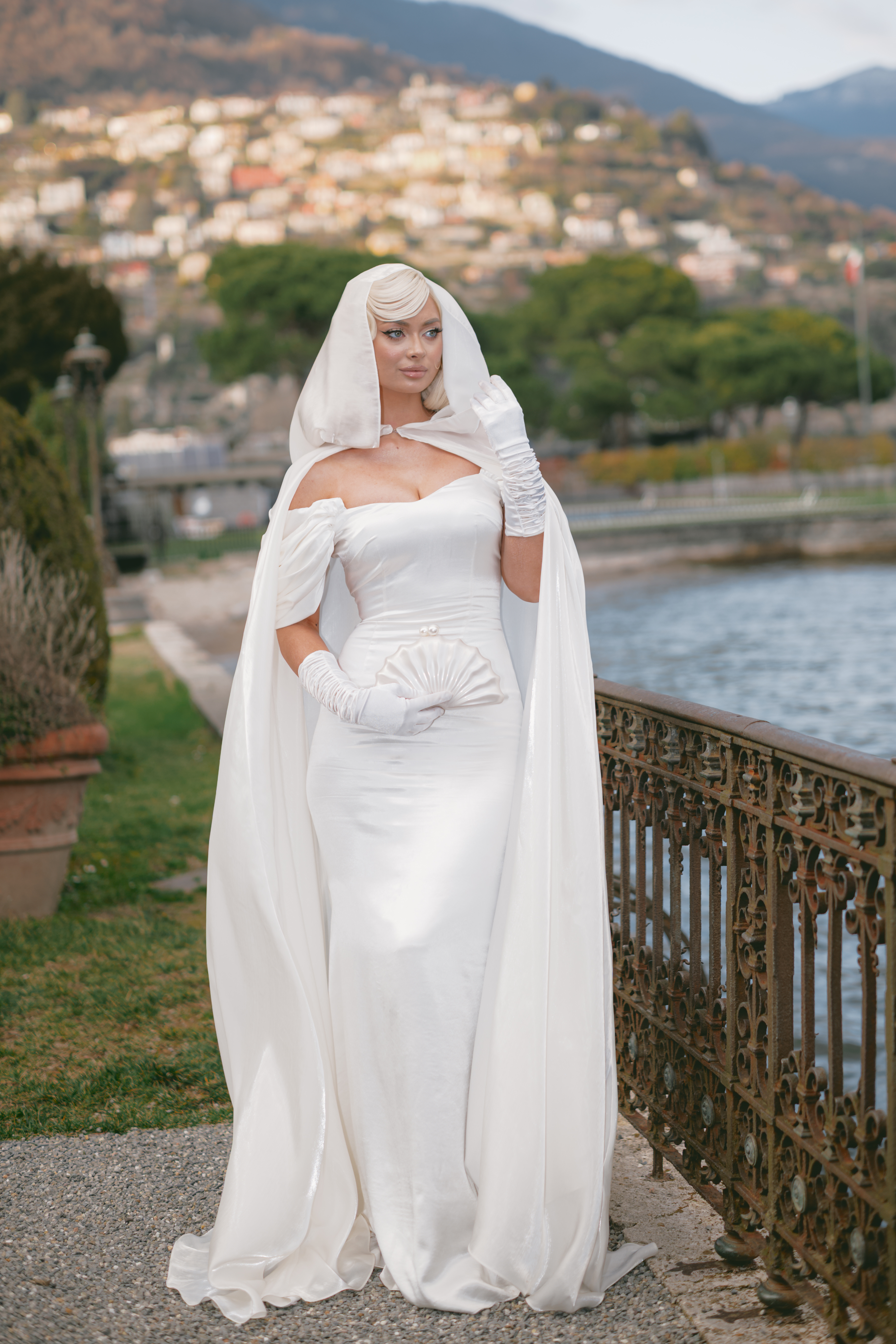 Woman in a white satin hooded cape and bridal gown wearing opera gloves by an ornate iron railing along a scenic lake.