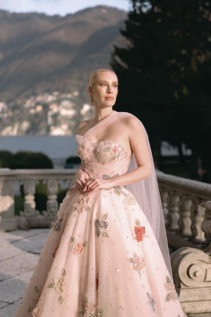 Woman in a pink floral embroidered gown standing on a stone terrace overlooking mountains and water