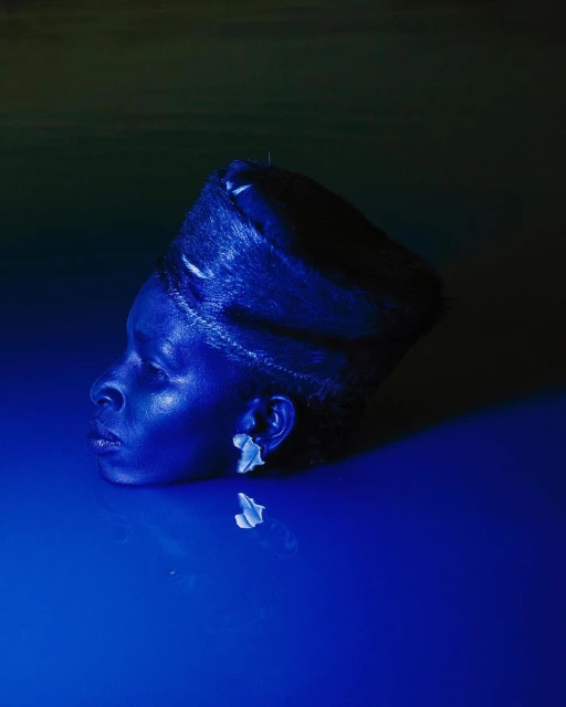 Portrait of a Black woman in water, wearing floral earrings and a head covering, bathed in blue light, creating a serene and artistic image.