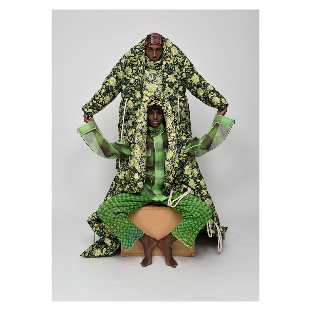 Two adult humans wear matching green floral outfits indoors in a studio shot; one is standing and the other is sitting on a suitcase.