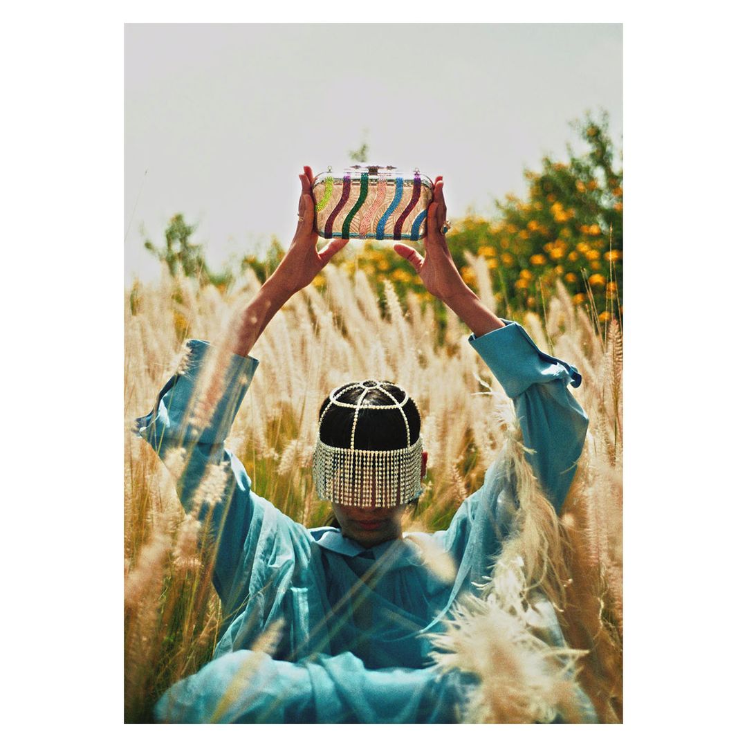 A woman in a rhinestone headpiece holds up a colorful striped handbag in a field of tall wheat-colored grass against a bright sky.