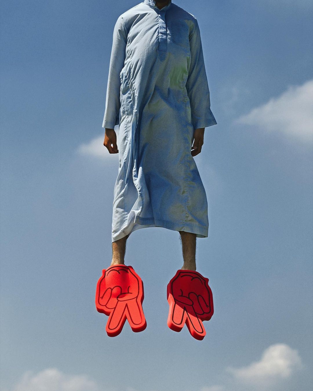 Man in traditional clothing floats in the sky wearing oversized red foam hands on his feet. Cumulus clouds and blue sky.