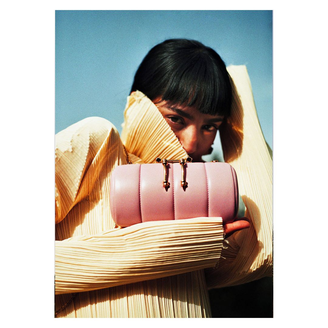 Woman presents a pink cylinder handbag in a fashion shot with a pleated top and blue sky, emphasizing accessory and stylish attire.