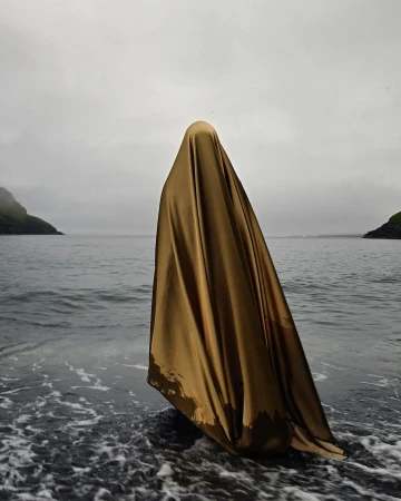 Figure draped completely in a shimmering gold fabric standing in calm water with rocky shores under an overcast sky