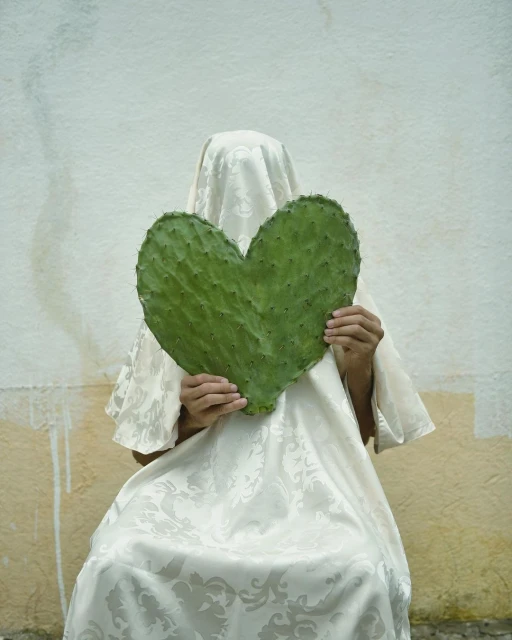 Person covered with patterned cloth holds a heart-shaped green cactus pad in front of a white wall, creating a surreal composition.