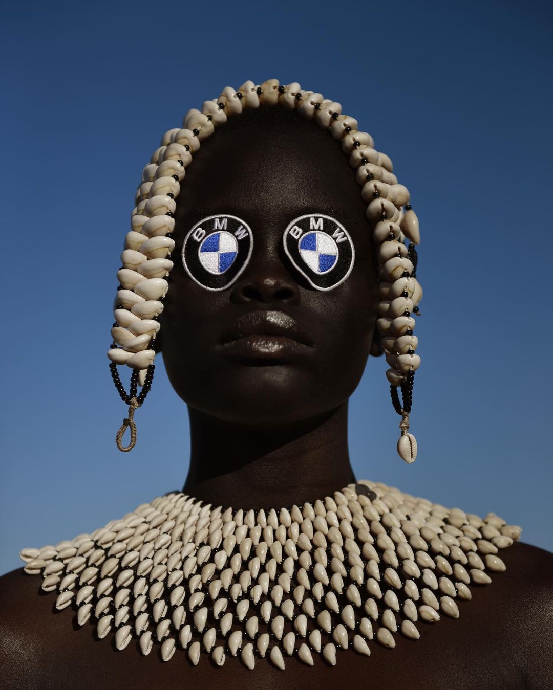 Woman with dark skin wears traditional shell necklace and headdress; BMW logos cover the eyes against a cloudless sky background.