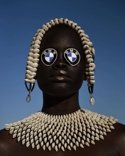 Woman with dark skin wears traditional shell necklace and headdress; BMW logos cover the eyes against a cloudless sky background.