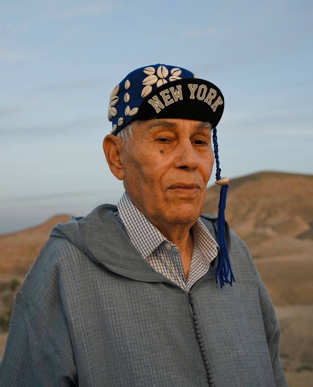 Elderly man in a blue robe and a 'New York' baseball cap decorated with shells posing in a desert landscape at dusk or dawn.