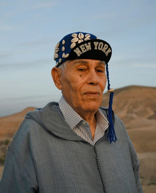 Elderly man in a blue robe and a 'New York' baseball cap decorated with shells posing in a desert landscape at dusk or dawn.