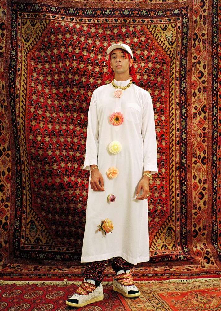 An individual with red hair wears a white tunic with flowers and patterned pants in front of an ornate patterned textile backdrop.