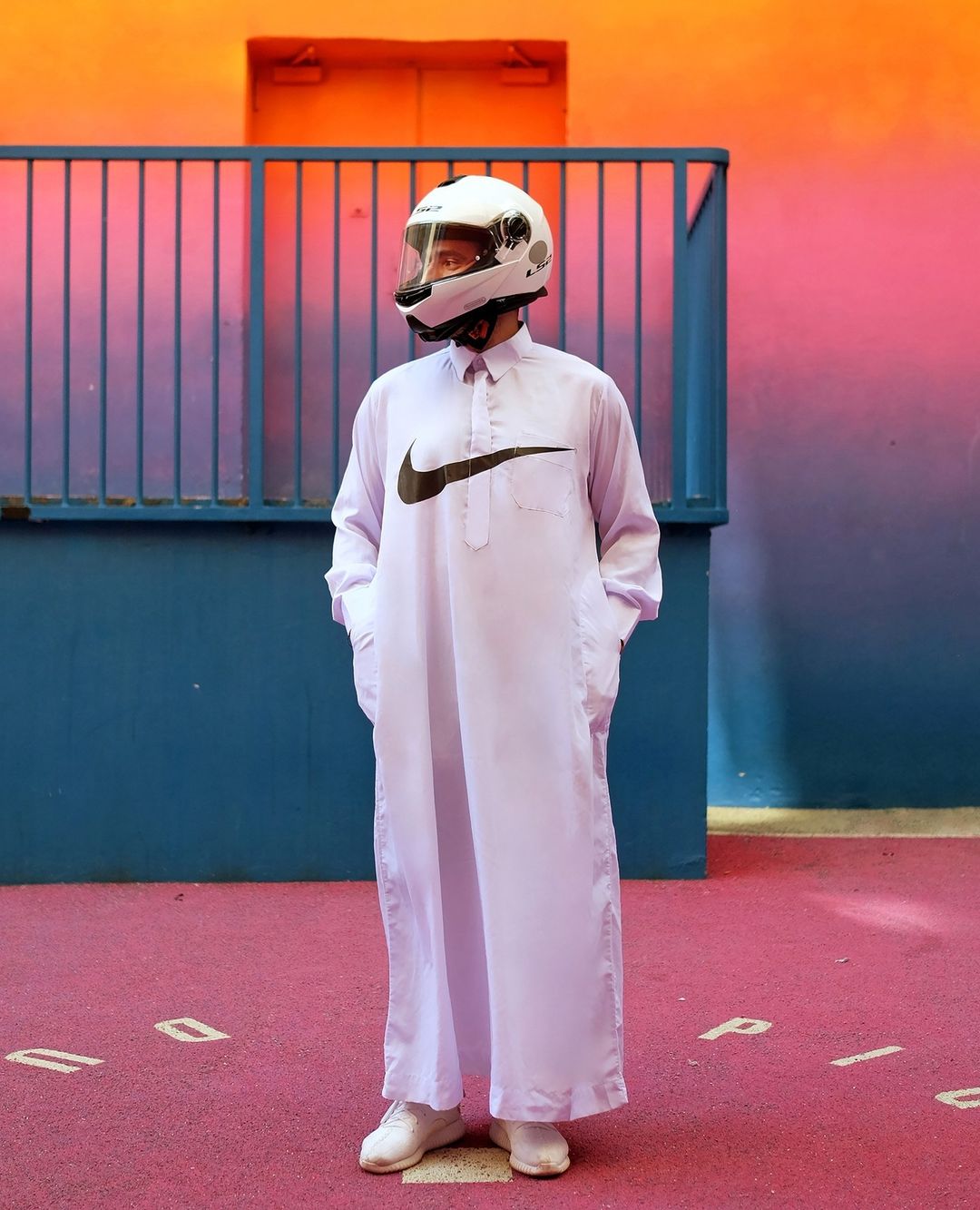 Adult male in a white thobe with a black Nike swoosh and white helmet stands against colorful pink, blue, and orange walls.