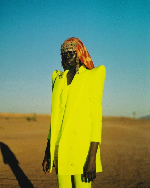 Adult woman in yellow suit wearing ornate headdress stands in desert landscape against blue sky in fashion-style outdoor portrait.