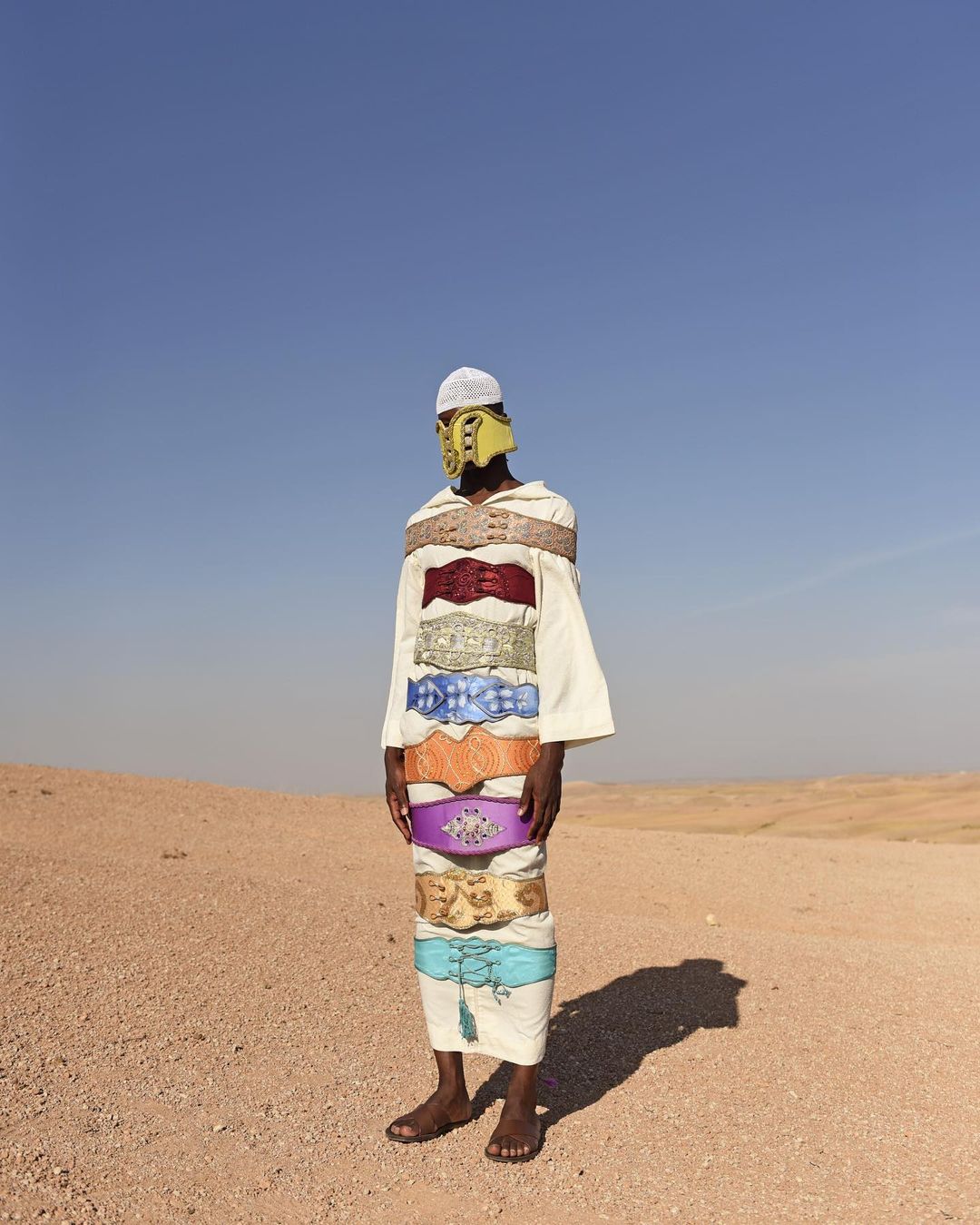 Person in desert landscape wearing white robe, headdress, yellow mask, and multiple colorful belts, creating a surreal fashion image.