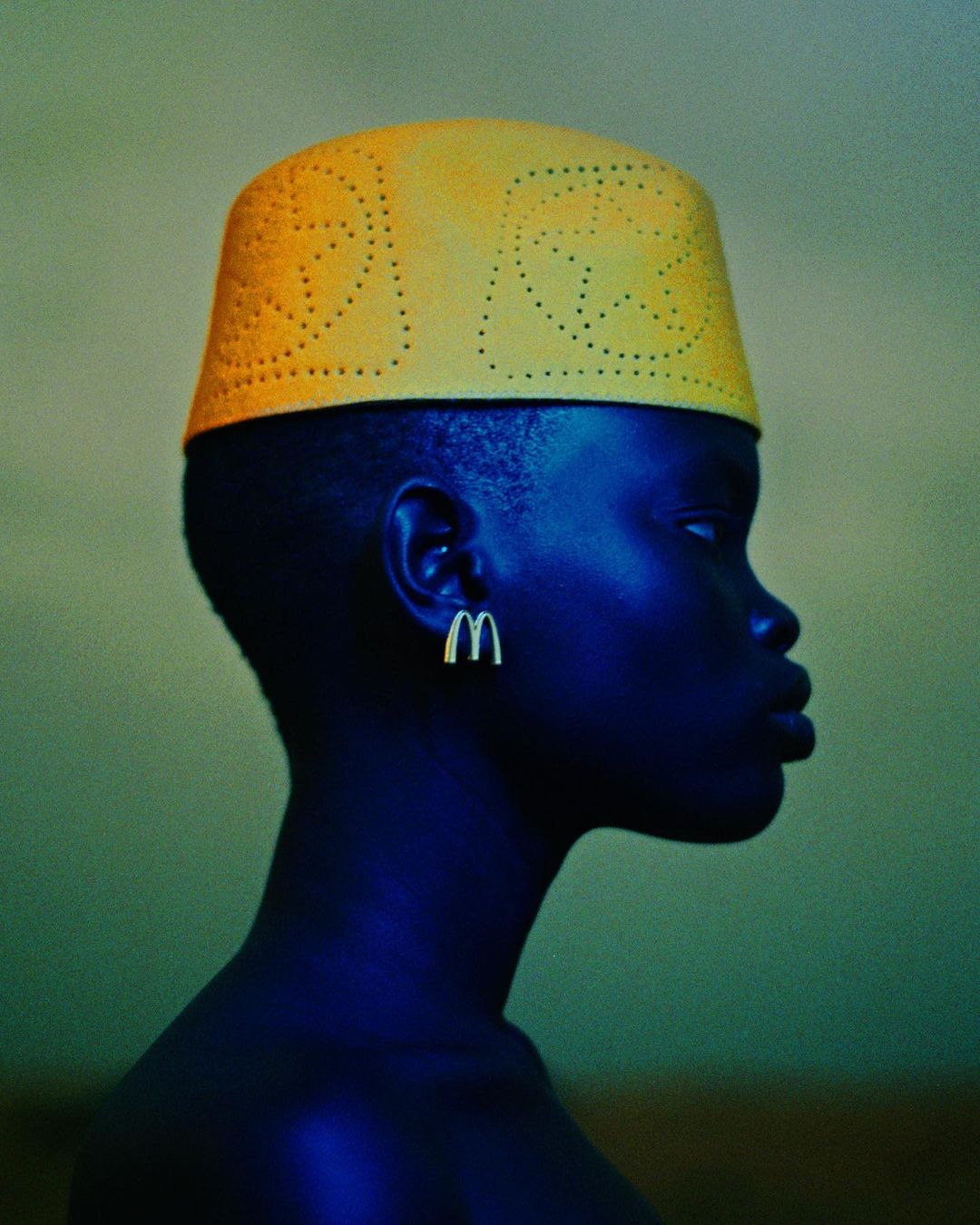 Profile portrait of dark-skinned woman wearing yellow hat and McDonald's logo earrings against muted green background. Studio shot.