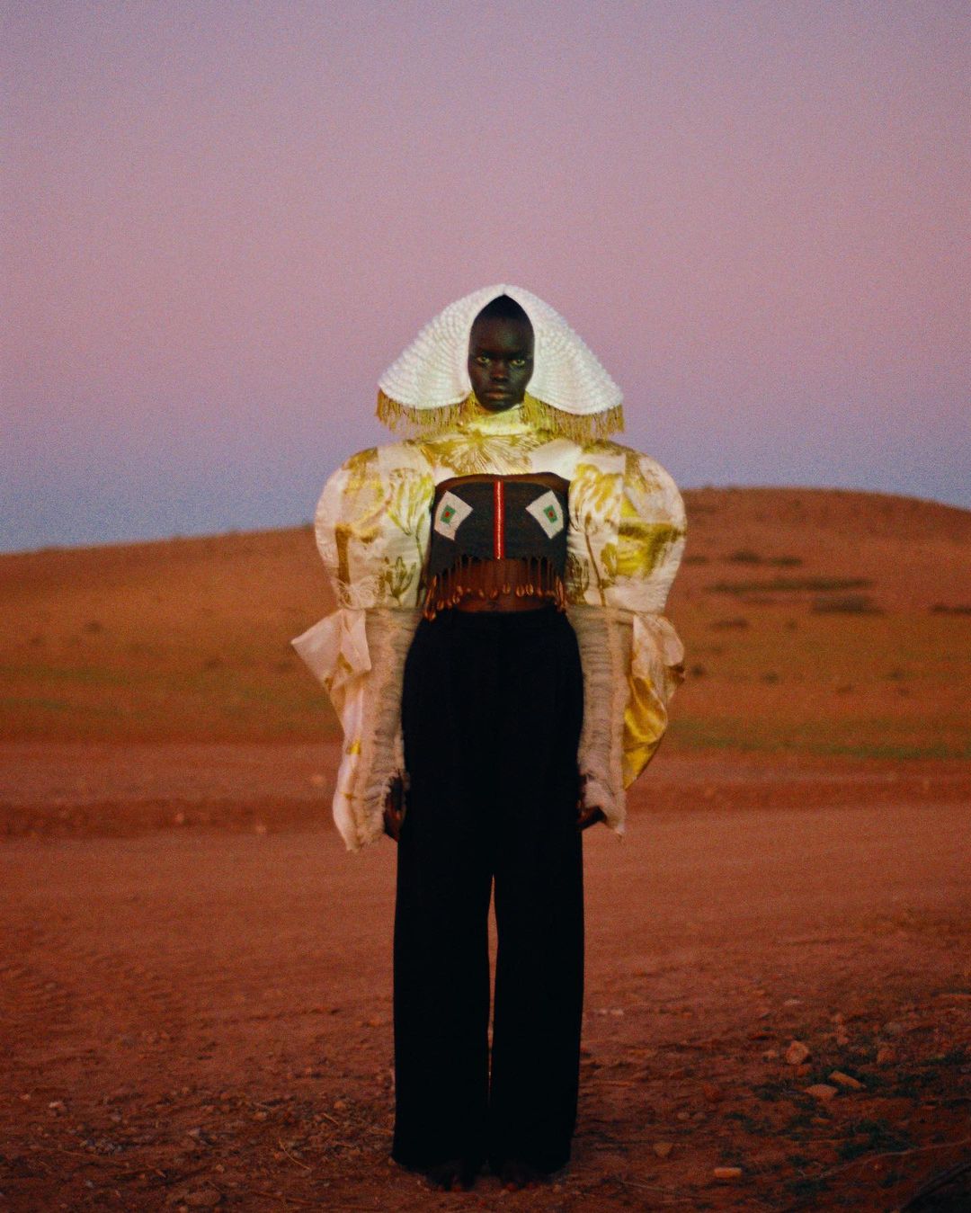 An African woman poses in a desert setting, wearing a custom couture outfit with a headpiece, statement sleeves, and black trousers.