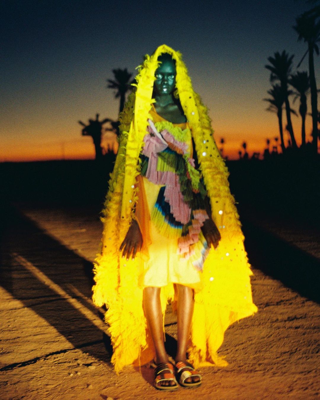 Fashion shot of an adult female with dark skin and a hooded yellow cape, posing outdoors against a sunset sky and palm trees.