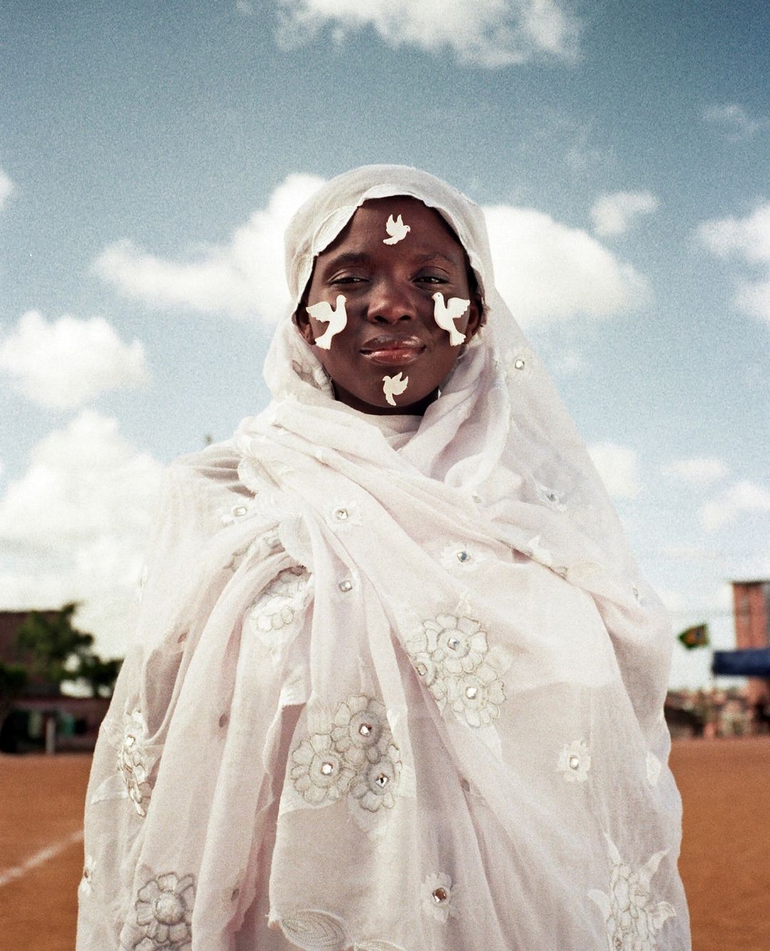Portrait of an African woman with white dove paintings on her face, wearing a white veil with floral embroidery outdoors.