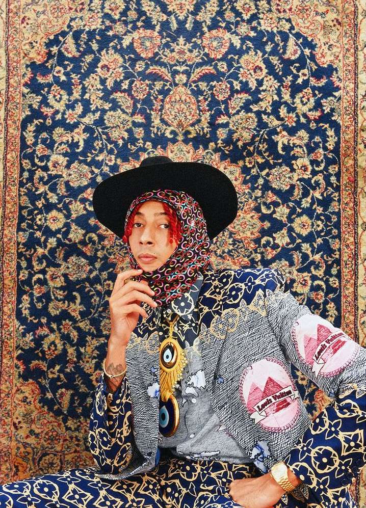 Man in patterned jacket, hat and headscarf poses in front of blue patterned textile backdrop, jewelry, fashion portrait.