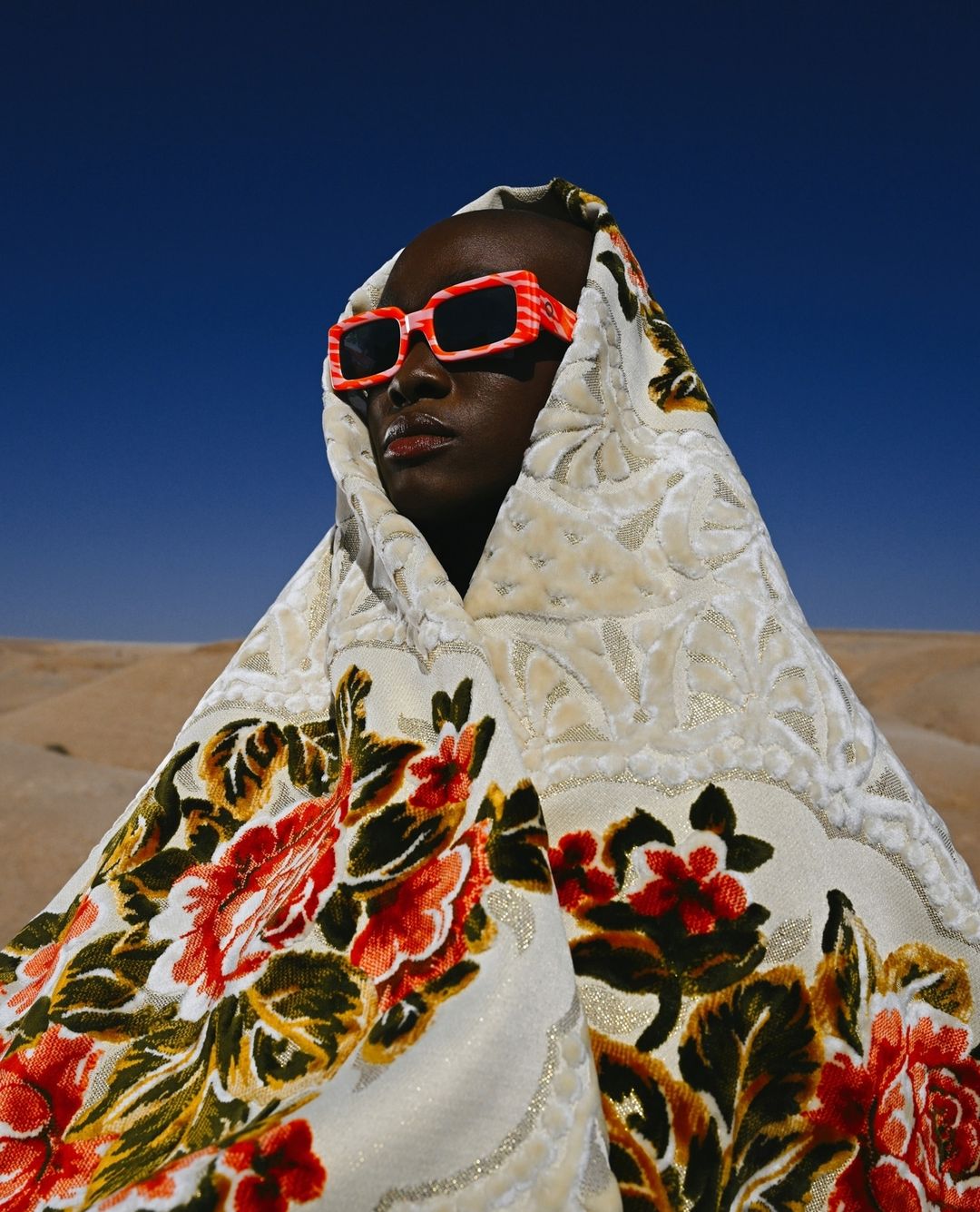 Person in floral-patterned head covering and red and white sunglasses posing in a desert setting with a blue sky background.