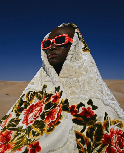 Person in floral-patterned head covering and red and white sunglasses posing in a desert setting with a blue sky background.