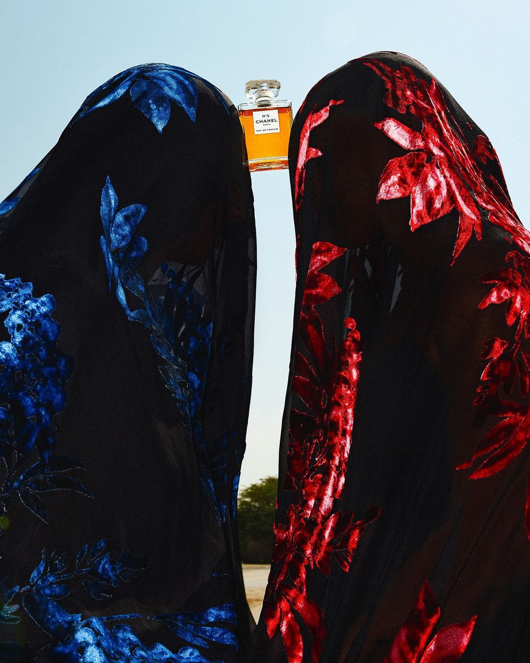 Fashion shot with two figures draped in black floral-patterned fabric flanking a perfume bottle against a pale sky backdrop.
