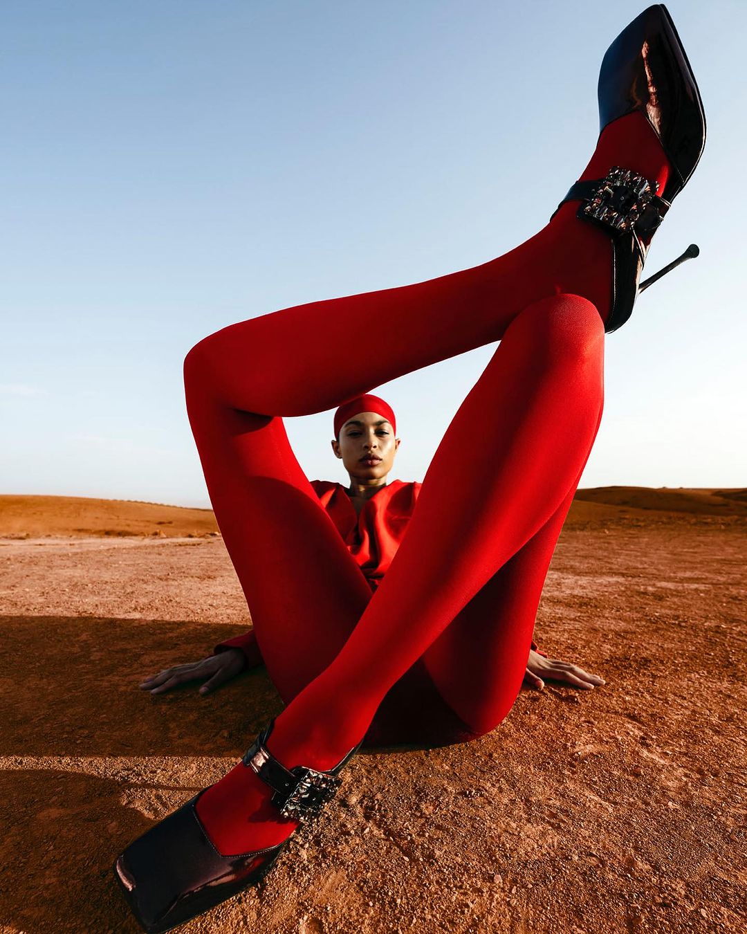 Fashion shot of a woman in red tights and heels sitting on a sandy surface against a blue sky, legs raised vertically.