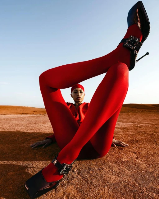 Fashion shot of a woman in red tights and heels sitting on a sandy surface against a blue sky, legs raised vertically.