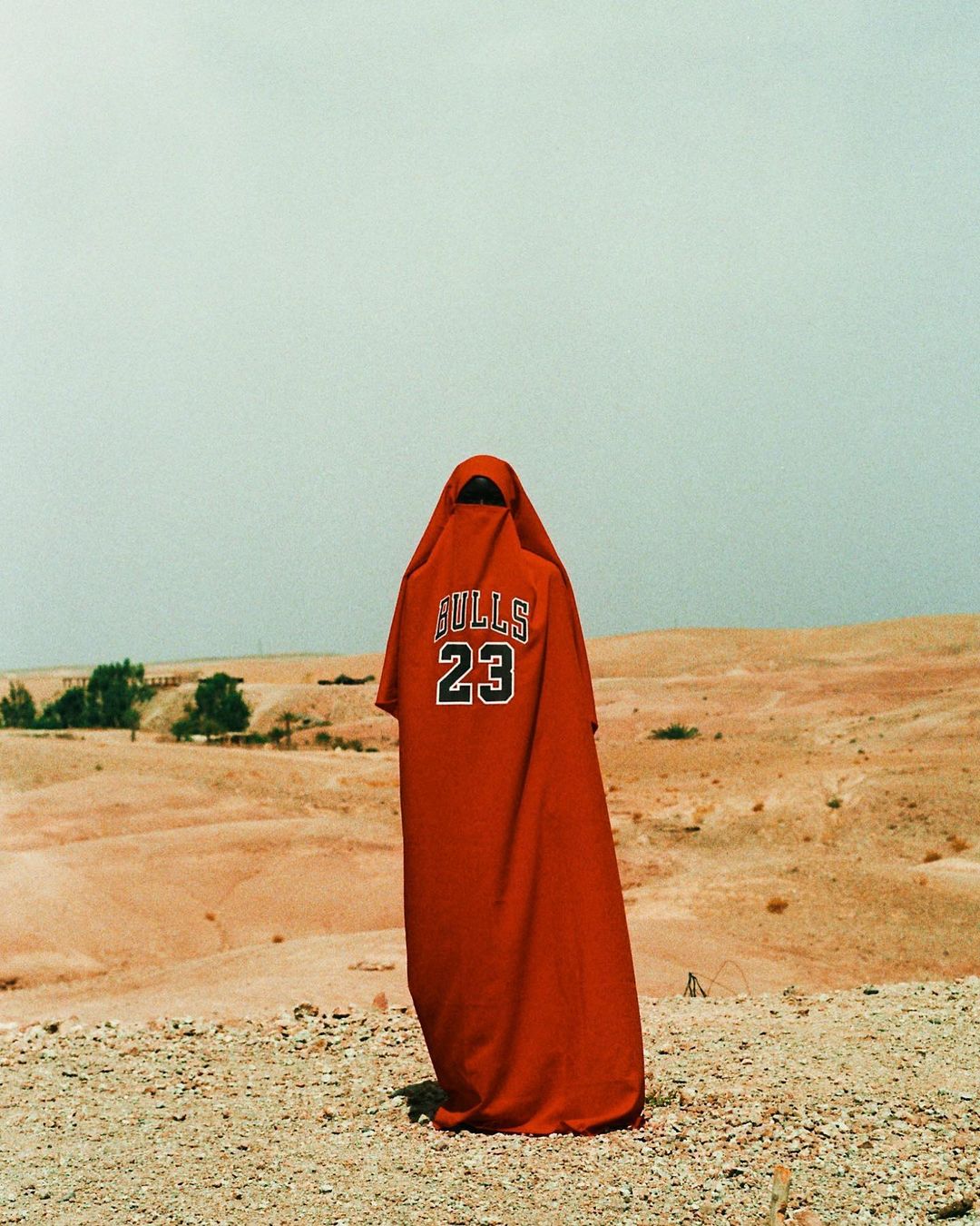 A figure in a red Bulls jersey-dress stands in a desert landscape under a bright sky, facing away from the viewer, enigmatic and bold.