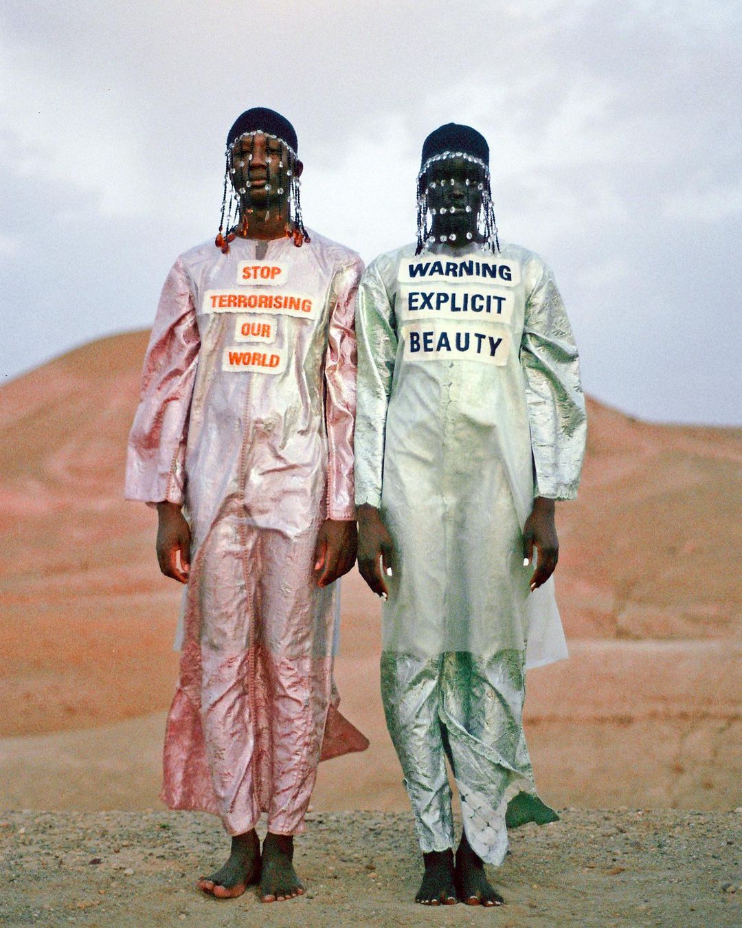 Two dark-skinned individuals in iridescent robes stand in a desert landscape, adorned with signage conveying social messages in an artistic manner.