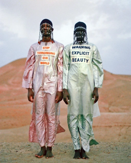 Two dark-skinned individuals in iridescent robes stand in a desert landscape, adorned with signage conveying social messages in an artistic manner.