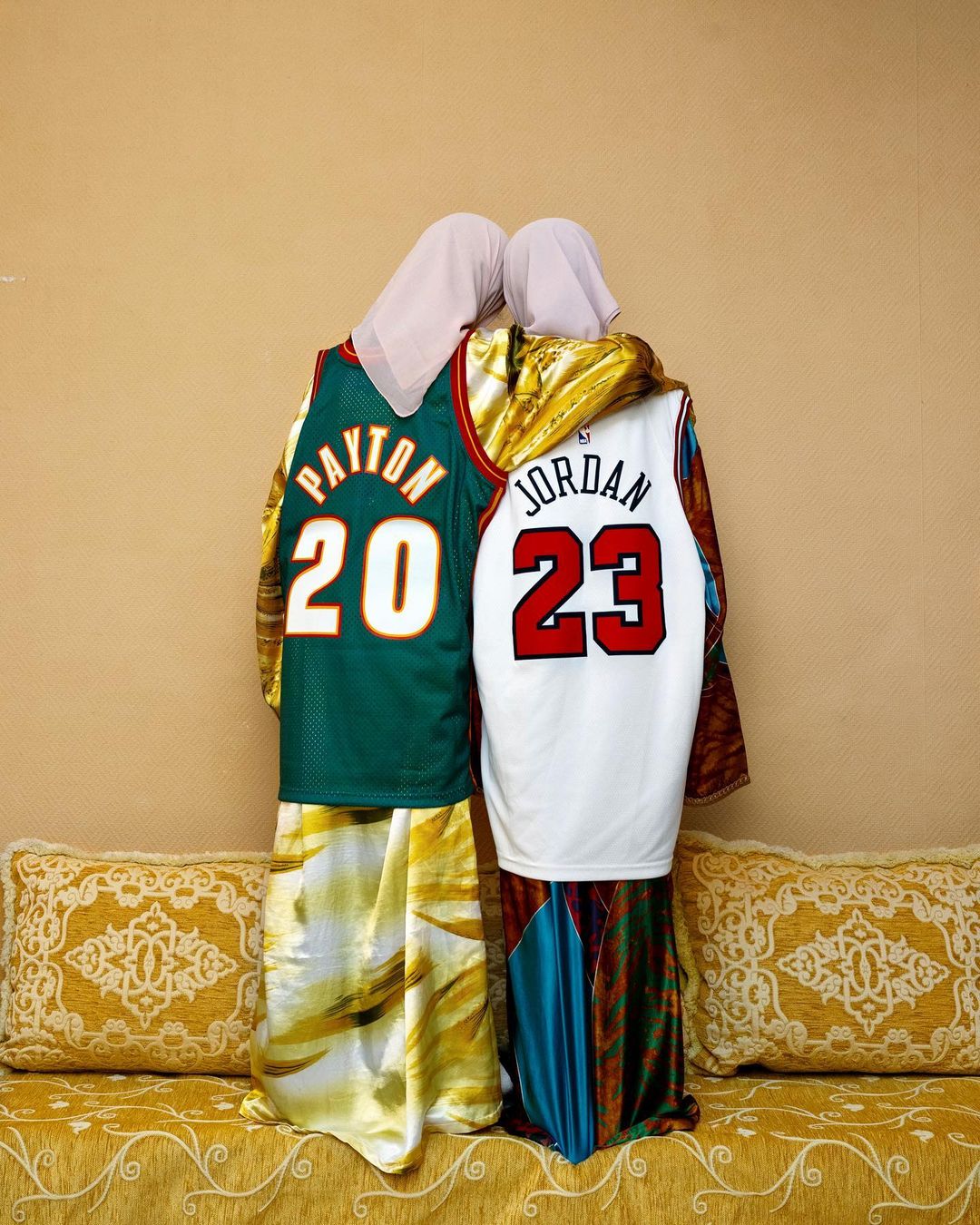 Two people in headscarves embrace, wearing vintage Gary Payton Sonics and Michael Jordan Bulls basketball jerseys, in an indoor setting.