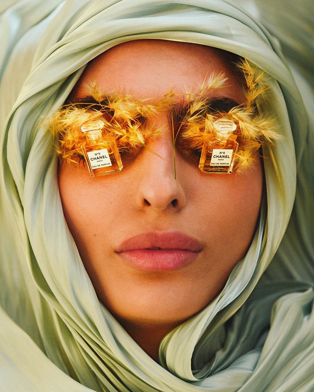 Conceptual beauty shot featuring a woman's face partially veiled in light green fabric, with Chanel perfume bottles and wheat in place of eyelashes.