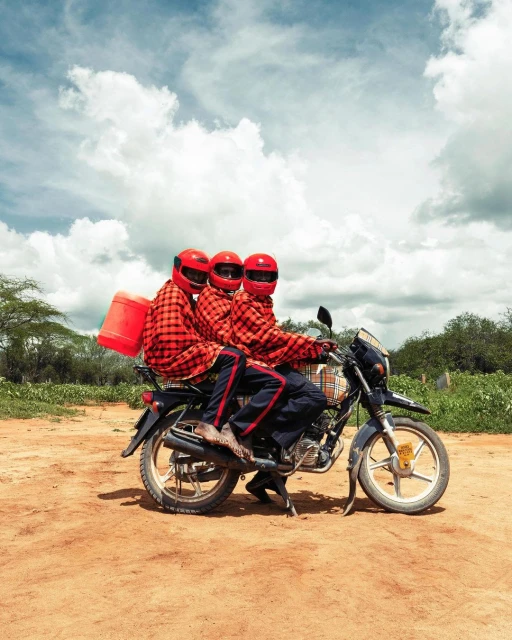 Three people wearing red plaid shirts and helmets ride a black motorcycle on a dirt road under a cloudy sky in an outdoor setting.