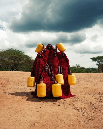 Group of six people in red robes carry yellow jugs in an arid, outdoor landscape under a cloudy sky, creating a striking, cultural image.