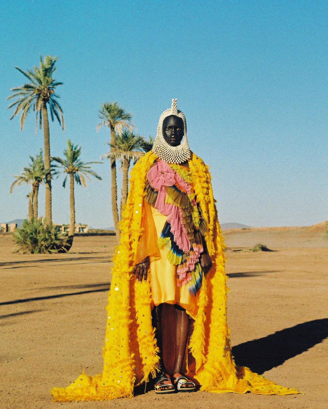 Person wearing a yellow fringed cape and beaded headpiece stands in a desert landscape with palm trees under a clear blue sky.