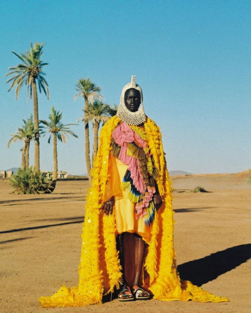 Person wearing a yellow fringed cape and beaded headpiece stands in a desert landscape with palm trees under a clear blue sky.