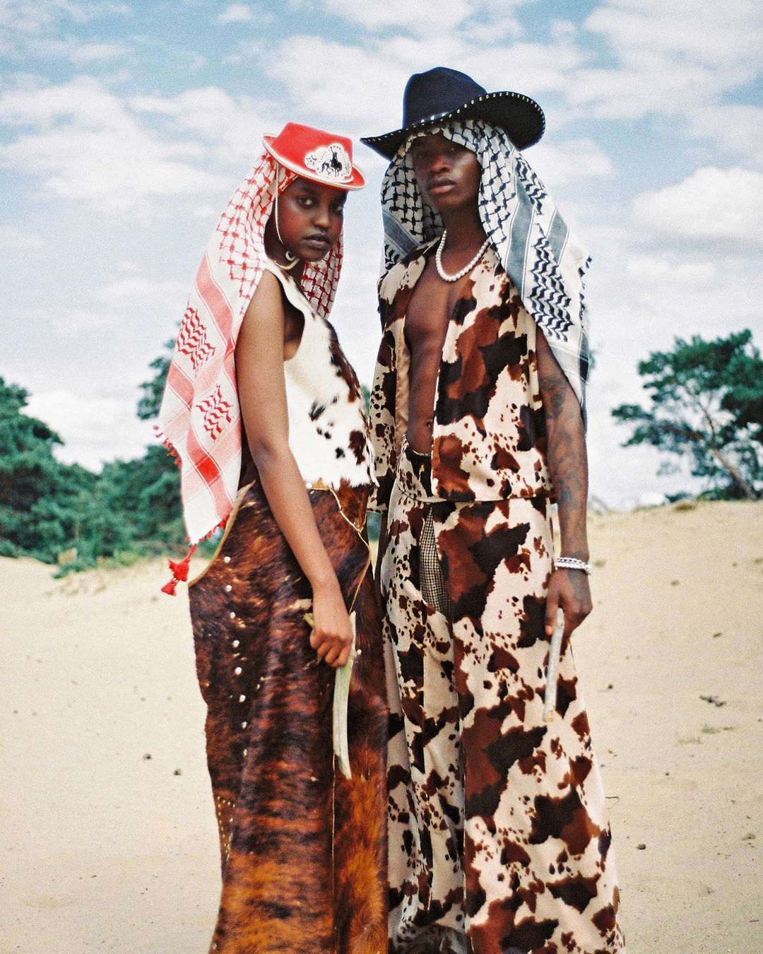 Two people in cow-print outfits and headscarves pose on a sandy landscape with scattered clouds and green trees in the background.