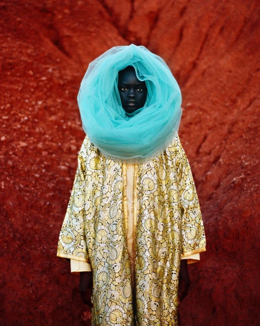 Person in patterned robe and turquoise veil stands before a textured red backdrop in a portrait capturing cultural fashion.