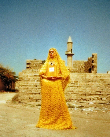 A woman in a yellow gown holds a Chanel perfume bottle, standing in front of an old brick building and minaret under a clear sky.