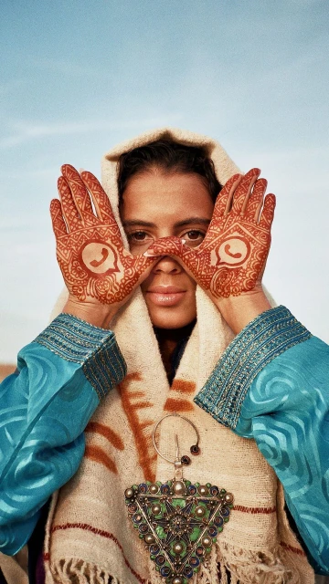 Woman with henna tattoos on hands and arms poses outdoors wearing a hooded garment and decorative pendant under a blue sky.