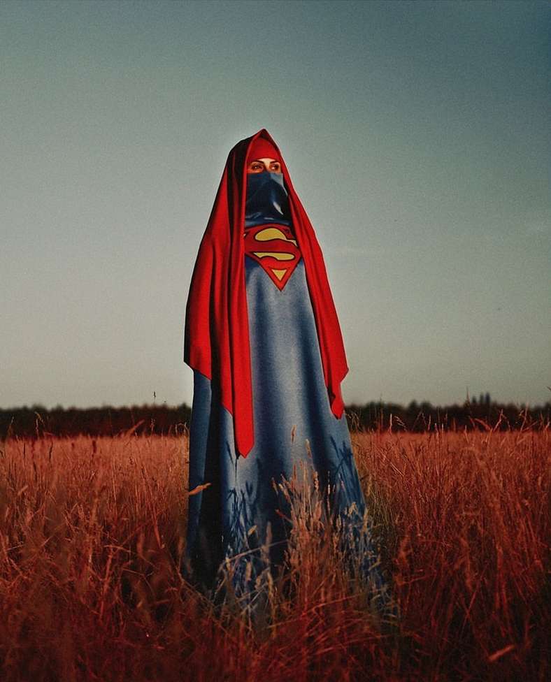 Person in a red cape and headdress stands in a field of wheat, evoking a powerful and striking visual statement with a pop-art style.