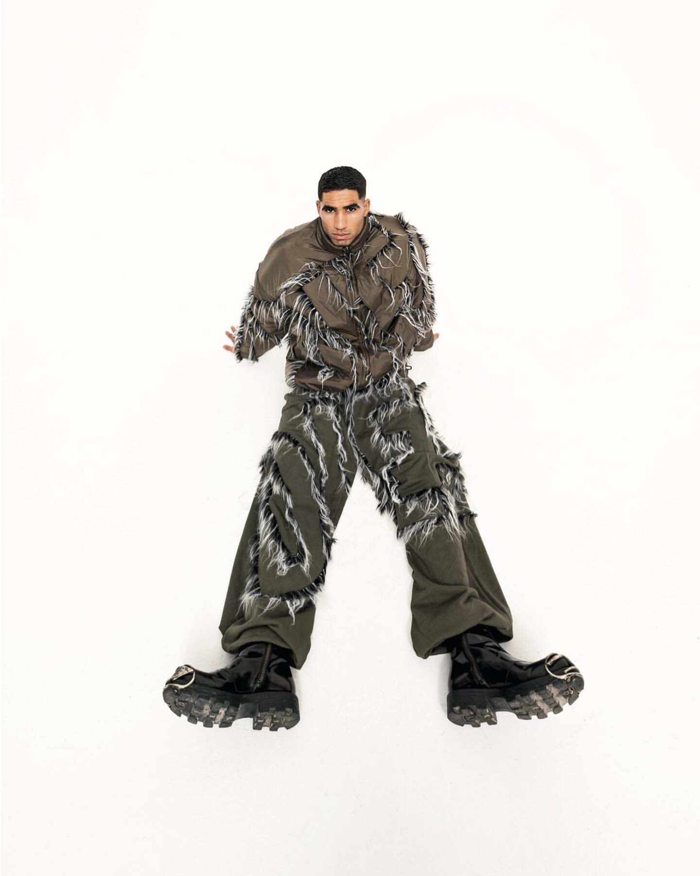 Fashion shot of a young adult man in a distressed brown jacket and green pants lying on a white surface with dark, shiny boots.