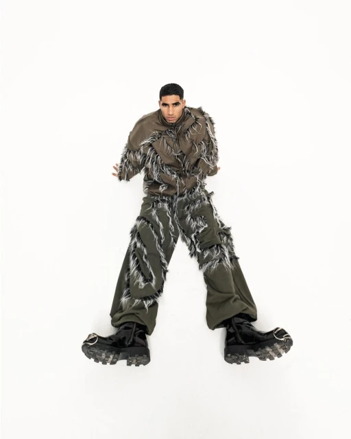Fashion shot of a young adult man in a distressed brown jacket and green pants lying on a white surface with dark, shiny boots.