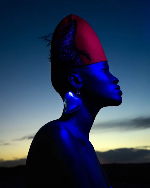 Profile of an adult female wearing a red fez, heart-shaped earrings, and illuminated with blue light against a sunset gradient sky.