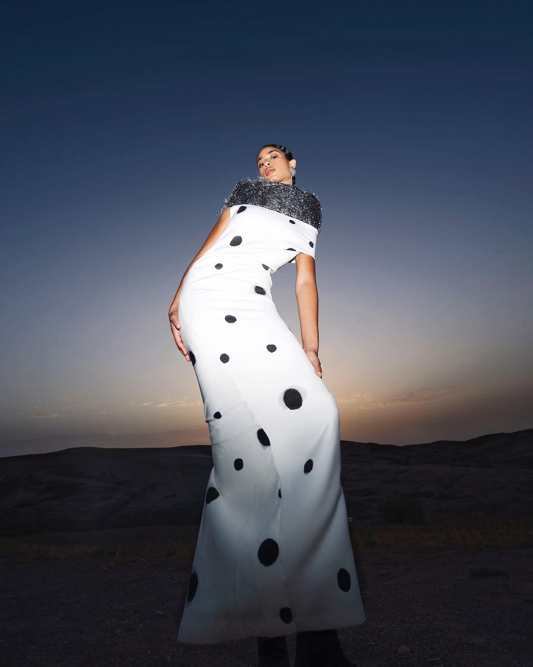 Woman in a white gown with black polka dots poses in a desert landscape at dusk, showcasing a unique blend of fashion and nature.