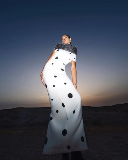 Woman in a white gown with black polka dots poses in a desert landscape at dusk, showcasing a unique blend of fashion and nature.
